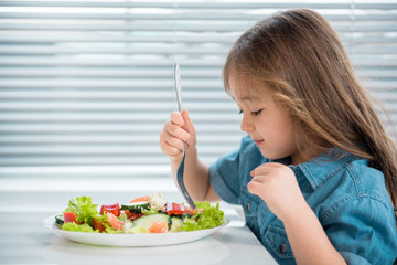 Profile of cute little asian girl having healthy breakfast at home. She is looking in plate of salad with interest while holding fork. Copy space