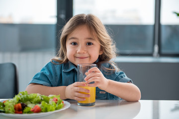 Waist up portrait of happy asian girl drinking fresh orange juice in the kitchen. She is looking at camera and smiling