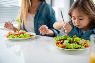 Cheerful mother and daughter are eating healthy salad together at home. Focus on plate with chopped vegetables
