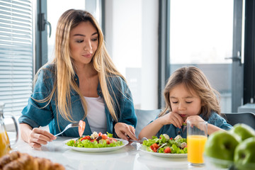 Pretty girl is looking at salad in plate with unwillingness. Her mother is giving fork to her while...