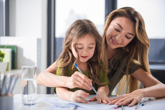 Wait up portrait of joyful mother and daughter drawing image together. They are sitting at home and laughing