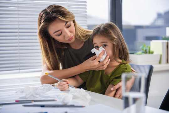 Asian Woman Is Taking Care Of Her Child. She Is Wiping Her Nose By Napkin With Gentleness. Girl Is Sitting At Table And Holding Pencil