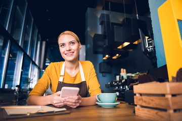 Fototapeta premium Low angle portrait of glad girl barista typing in phone while tasting cup of coffee at table in confectionary shop. Rest concept