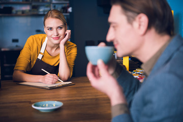 Portrait of cheerful young female barista watching at client while noting information. He tasting appetizing cup of beverage in confectionary shop. Job concept