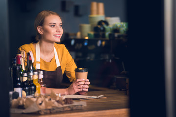 Portrait of beaming worker tasting mug of beverage while resting elbow on counter in confectionary shop. Break concept