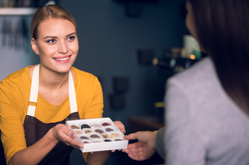 Portrait of beaming female pastry chef keeping delicious sweets in arms. She showing them to visitor in chocolate house. Work concept