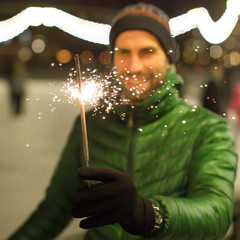 Happy man with beard holds Bengal fires, smiles and looks at the sparks outdoors/focus on sparks, ice rink and lights on background
