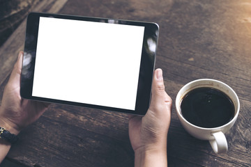 Mockup image of business woman's hands holding black tablet pc with blank white screen and coffee cup on vintage wooden table in cafe background