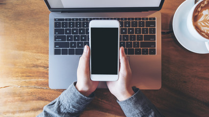 Top view mockup image of business woman holding mobile phone with blank white screen with laptop on wooden table in cafe