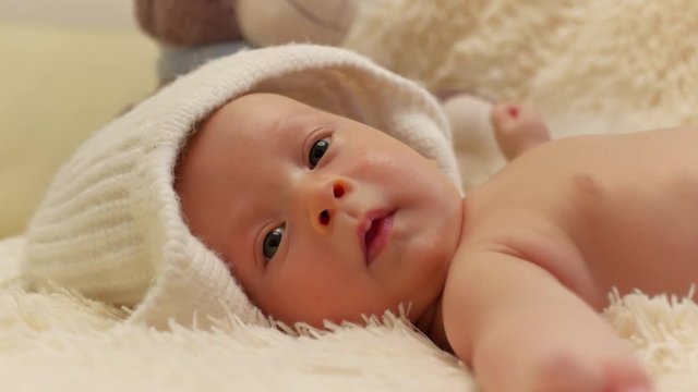 A charming little baby is looking into the camera and is wearing a white hat.