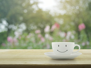 White coffee cup with smile face shape on wooden table over blur pink flower and tree in garden, soft tone, Good morning concept