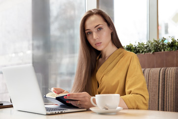 beautiful stylish long-haired woman is planning your day thumbing through the diary sitting at a table in the cafe with a laptop and a Cup of tea