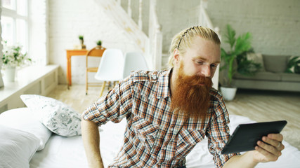 Young bearded man using tablet computer having video chat sitting in bed at home