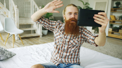 Young bearded man using tablet computer having video chat sitting in bed at home