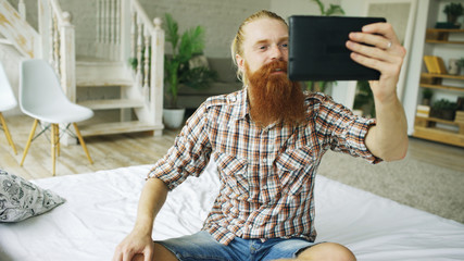 Young bearded man using tablet computer having video chat sitting in bed at home