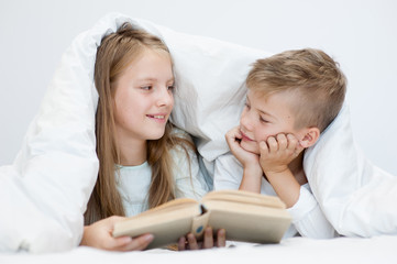 Two happy sibling children lying under blanket with book