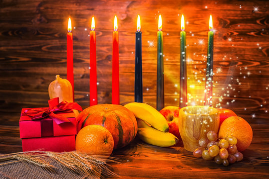 Kwanzaa holiday concept with traditional lit candles, gift box, pumpkins, ears of wheat and miracle magic light on wood background