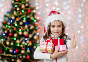 Happy little girl holding many gift boxes and showing thumbs up.Space for text