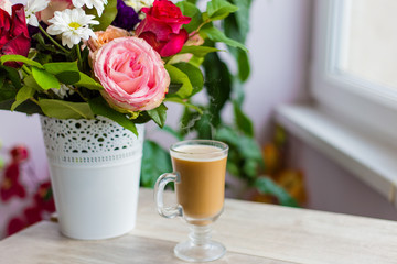 cup of hot coffee with milk stands near the window and a bright colored bouquet of flowers
