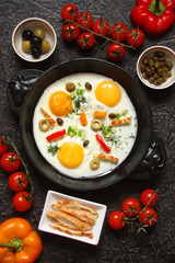 Fried eggs in a cast iron pan with peppers, tomatoes, capers, herbs and bread on a black background