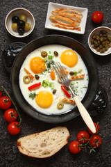Fried eggs in a cast iron pan with peppers, tomatoes, capers, herbs and bread on a black background