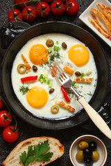 Fried eggs in a cast iron pan with peppers, tomatoes, capers, herbs and bread on a black background
