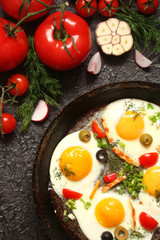 Fried eggs in a cast iron pan with peppers, tomatoes, capers, herbs and bread on a black background