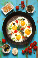 Fried eggs in a cast iron pan with peppers, tomatoes, capers, herbs and bread on the blue boards