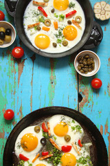 Fried eggs in a cast iron pan with peppers, tomatoes, capers, herbs and bread on the blue boards