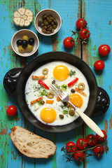 Fried eggs in a cast iron pan with peppers, tomatoes, capers, herbs and bread on the blue boards