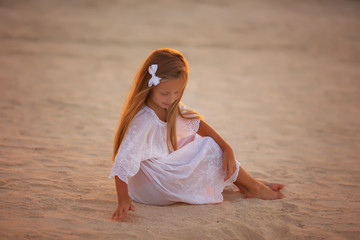 Fototapeta na wymiar Little cute girl with long hair in a white tunic playing with sand on the beach at sunset