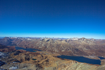 From left to right of the viewpoint, the Lake Sils and Lake Silvaplana are in dark blue under the clear blue sky. The lakes are located close to St. Moritz of Switzerland.