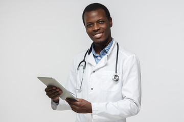 Handsome african doctor standing isolated in grey background holding tablet device, showing confidence and optimism while giving straight open look and smiling