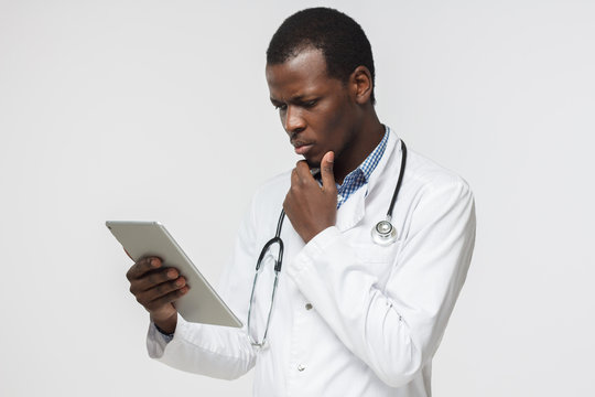 Picture of young african doctor thinking about treatment, holding tablet PC while using medical app isolated on grey background