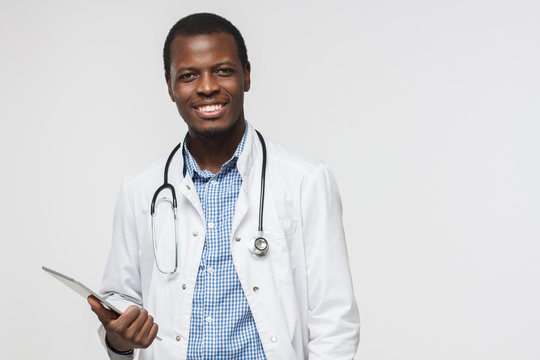 Horizontal photo of african doctor isolated on grey background smiling happily and holding his tablet computer. Concept of contemporary gadgets and medical services