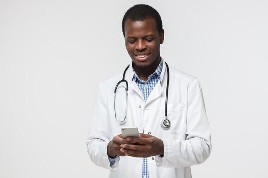 Closeup photo of african doctor standing isolated on gray background looking attentively at screen of cellphone, smiling nicely