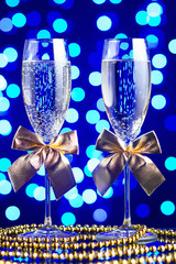 Festive glasses with champagne, golden bows and beads on a glass table with a beautiful blue bokeh - 184143223