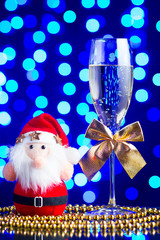 Festive glass with champagne, golden bows, beads and Santa Claus toy on a glass table with a beautiful blue bokeh - 184143238