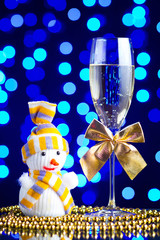 Festive glass with champagne, golden bows, beads and Snowman toy on a glass table with a beautiful blue bokeh - 184143261