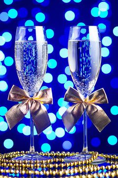 Festive glasses with champagne, golden bows and beads on a glass table with a beautiful blue bokeh
