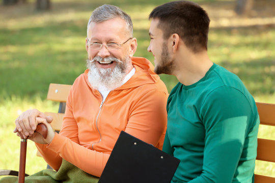 Young caregiver sitting with senior man on bench in park