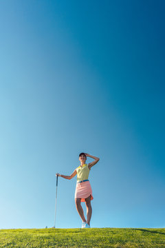 Low-angle view of a fit and cheerful woman wearing golf trendy outfits, while looking away during practice on the green grass of a professional golf course