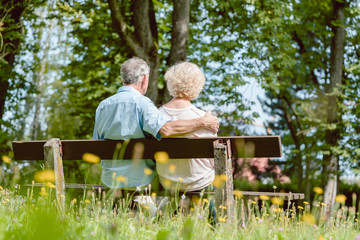 Rear view of a romantic elderly couple enjoying nature while sitting together on a bench in a...