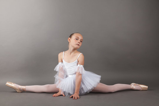 Young beautiful child girl ballerina doing split practicing ballet wearing white tutu dress on light gray background. Samakonasana stretching exercise, straight angle pose.