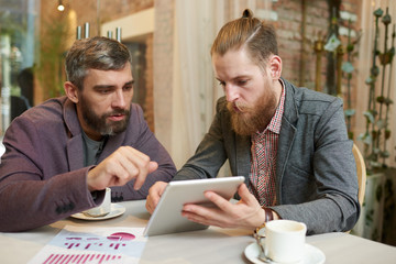 Portrait of two modern business people discussing work and using digital tablet during meeting in cafe on coffee break