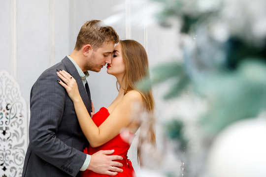 Young couple kiss in evening outfits (jacket and red dress) in white christmas interior - Powered by Adobe