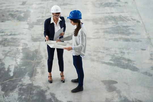 Confident businesswoman wearing hardhat studying blueprint with help of pretty young industrial engineer while carrying out inspection at production department of modern plant