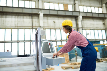 Profile view of confident young worker wearing hardhat and overall operating machine while standing at spacious industrial workshop, portrait shot