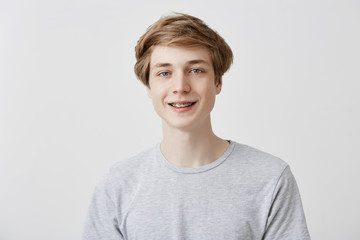 Happiness and positive human expressions. Studio shot of friendly young male student with fair hair and blue eyes laughing at good joke, smiling with braces, looking at camera with carefree smile