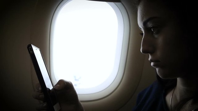 Young gir is using a smartphone inside an airplane next to a window